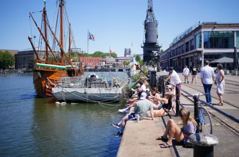 People sit on the edge of Bristol Harbour on a sunny day. In the background The Matthew ship and the MShed museum are visible
