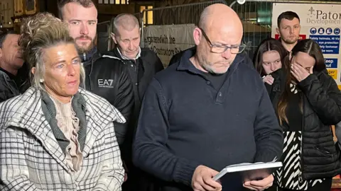 David Knight, the uncle of Mason Rist, reads out a statement on a notepad outside of Bristol Crown Court. On his right is Mason's mum, Nikki Knight, who stands beside him with a sad expression. The people around him have sad expressions and one girl is in tears