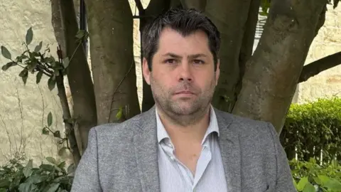 Leigh Frost is looking serious and at the camera. He is standing in a garden, and there is a tree and hedging behind him. He has dark hair and a slight beard. He is wearing a grey jacket over a grey and white striped shirt.