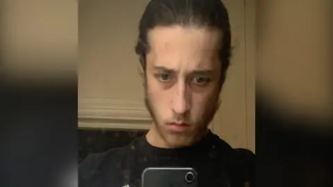 Met Police Alfie Coleman looking serious with his dark hair pulled backwards. This is a selfie and the top of his iPhone is pictured.