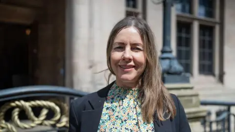 Staffordshire County Council A woman with brown hair stands in front of a large building. She is wearing a floral blouse and a blazer.
