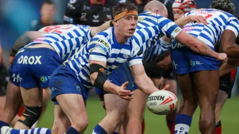 Several rugby players wearing blue and white striped shirts, blue shorts and blue and white striped socks during a match. The player at the centre of the screen is passing the ball to a teammate out of the frame.