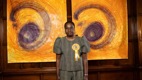 PA Media A woman with short black hair wearing a matching checked top and trousers and a yellow rosette stands in an art gallery. She is standing in front of two large yellow spiral drawings. 