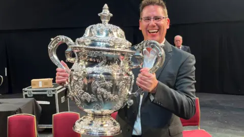 Flowers Band  Paul Holland smiling to camera holding the enormous silver trophy in his arms