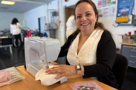 A women wearing a white short and black cardigan uses a sewing machine while sitting at a wooden desk