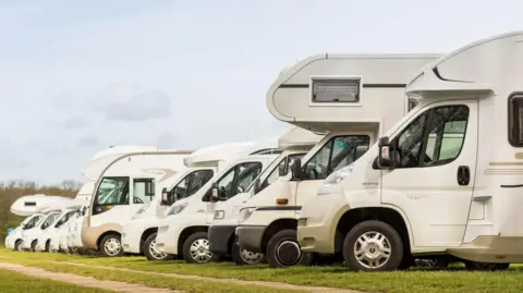 Merten Snijders/Getty Images Side view of a row of motorhomes of varying size and design parked on a grass field with trees in the background