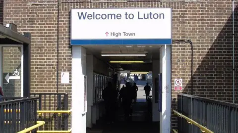 The outside of Luton Railway station, showing a large white sign, people walking into the building, railways and a brick building. 