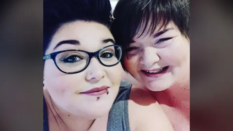 Lizzie Lyle This is a selfie of two women - Lizzie Lyle and her mother Alison McKinney - both smiling at the camera. Ms Lyle, on the left, has dark hair brushed off her face. She is wearing dark-framed glasses and has two lip piercings. Ms McKinney on the right is smiling and has a dark fringe brushed forward.