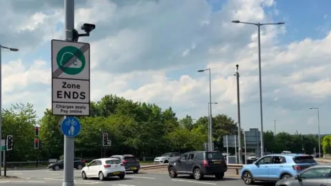 A busy urban road junction on a partly cloudy day. In the foreground, there is a tall grey pole positioned slightly to the left of the center. Attached to this pole is a rectangular road sign with a green circular symbol that contains a cloud-like shape and the letter “C” A diagonal black line crosses through the symbol, indicating the end of a designated zone. Beneath the symbol, the sign clearly reads “Zone ENDS” in bold black letters, followed by smaller text stating “Charges apply Pay online”. Below this sign, there is a smaller circular blue sign showing a white bicycle and a pedestrian, indicating a shared path for cyclists and pedestrians.