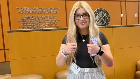Madi Foster A woman with blonde hair stood in front of a brown wall and panels with signs and logos. She is holding two thumbs up to the camera, with her phone in one hand. The writing on the wall and accompanying test refers to The National Institutes of Health.