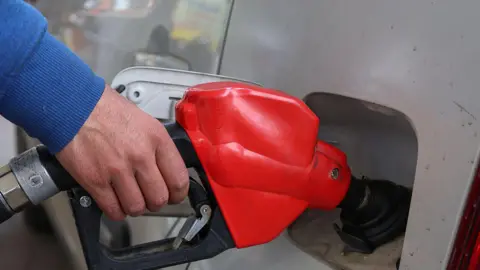 A close-up image of a silver car being filled up at a fuel pump, the pump is red and black. The person holding the pump is wearing a blue jumper. 