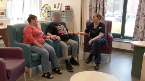 The photo shows a small lounge area inside a care or medical facility. Three people are seated on armchairs arranged in a semi-circle: two are sitting together on a sofa, while another, dressed in a navy uniform, sits opposite them holding one person’s hand.
The room has light wood flooring, a round coffee table in the foreground, and large windows with patterned curtains letting in natural light. In the background, there are activity materials and a colourful game board on a shelf, suggesting a therapeutic or social setting.
