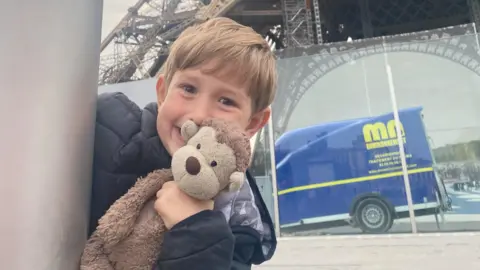 Family handout A young boy peeking round the corner of a grey post smiling in a cheeky way and holding a brown monkey teddy up to his cheek. He has dark blond hair and a fringe and is wearing a black coat. The bottom of the Eiffel Tower is in the background, as is a reflection of a blue van.