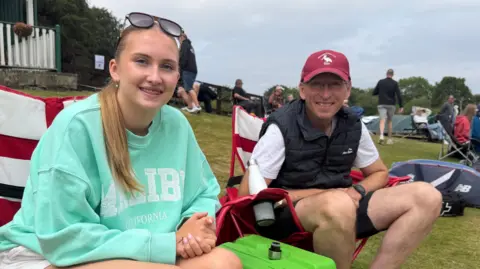 Martin and Poppy sitting on red and white camping chairs and smiling into the camera. Poppy is closest to the camera with light brown hair tied back in a long ponytail. She has sunglasses on top of her head and braces. She is wearing a turquoise jumper with writing on it and has her hands clasped in her lap. Her dad, Martin sits on the right and is hearing a red cap and black bodywarmer on top of a white t-shirt and shorts. There are people sitting around them on the grassy hill.