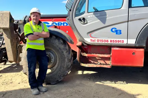 Construction worker in a high-visibility shirt and hard hat stands with arms crossed beside a large red and grey telehandler on a dusty worksite under a clear blue sky.