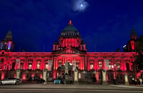 The front of a stone building with domes and lots of windows, lit up red against a night sky
