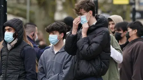 Three young men wearing jackets and blue medical face masks stand outside