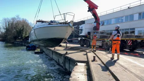 A boat is being hoisted out of the water by a crane with men in high visibility outfits holding rope attached to it and guiding it towards the shore.