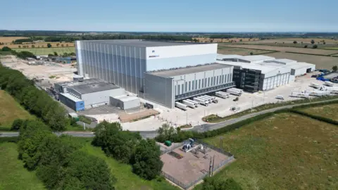 BBC/Joe Bilton The Magnavale plant - a large warehouse building clad in white blue and grey with a distribution point at the front for HGV trailers and several smeller buildings. It is set in green fields under a blue sky.