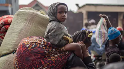 AFP A young boy sitting on an adult's shoulders stares at the camera as residents carry their belongings as they flee from Kibati, where fighting has intensified, towards the city of Goma on January 26, 2025.