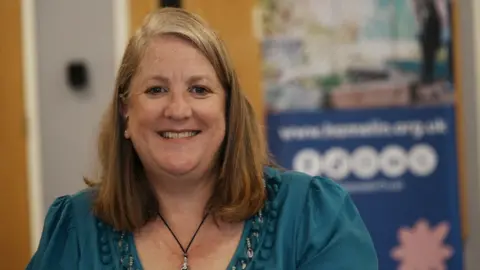 John Fairhall/BBC Shoenagh Mackay sits in front of a blue poster with writing on it. She is wearing a turquoise top with a black necklace. She has shoulder-length blonde hair.