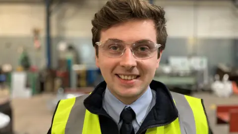 Will Underwood stands inside Underwoods' Worcester plant. Welding equipment is blurred in the background. Will is wearing a light blue shirt, navy blue tie and black fleece, as well as a hi-vis jacket and safety glasses.