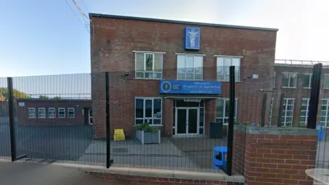 Google Maps Exterior view of brown school building with blue signs and tarmac courtyard, behind a black metal fence.