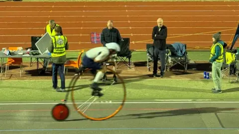 Rory Hitchens Richard Thoday on his orange framed penny farthing at Reading Velodrome. Behind him stood on the side of the track are three people wearing yellow hi-vis fluorescent vests and two men both dressed in black.