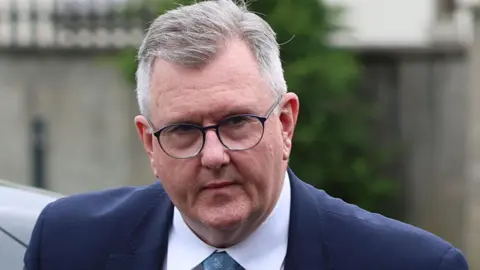 PA Media A man with short grey hair and glasses is seen in close up, with only his head and shoulders visible. He is wearing a blue suit jacket, white shirt and a turquoise patterned tie, the tops of all of which can just about be seen. He is outside, possibly walking away from a car he has just exited as the top of the dark-coloured car can be seen behind him.