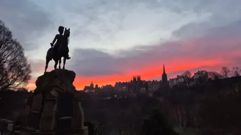 Robyn Campbell A pink sunrise and grey clouds over shadowy buildings in the distance. A statue of a person on a horse is in the foreground.