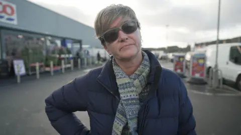 Fiona Macdonald, who is wearing a blue jacket, scarf and sunglasses, poses with her right hand on her hip outside Tesco on Stornoway