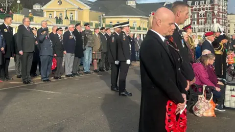 Crowds gathered in on Douglas Promenade with the heads bowed. Some people are in military dress or official uniforms and other are holding red poppy wreaths.