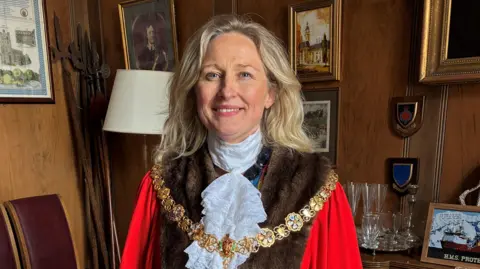 Cambridge City Council Maria Cleminson smiles at the camera. She has blonde hair, which falls just below the shoulders. She wears ceremonial mayoral robes, with a gold chain. She stands in a wood-panelled room with a variety of pictures and drinking glasses on display. 