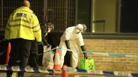 Police tape cordons off the scene, with two forensics officers picking things up from the floor.
