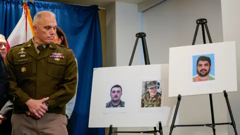 REUTERS/Nathan Howard A man in uniform in a press room with a flag and blue curtain behind him is at the left side of the frame looking at two poster boards propped up on easels. One poster board has two photos of the National Guard members who were shot and another has the photo of the suspect.