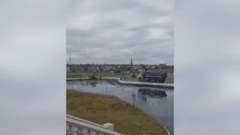 Alec Brown Clean up of Coatham boating lake. A lorry and an excavator are removing items from the nearly empty lake. There is a row of houses in the background. 
