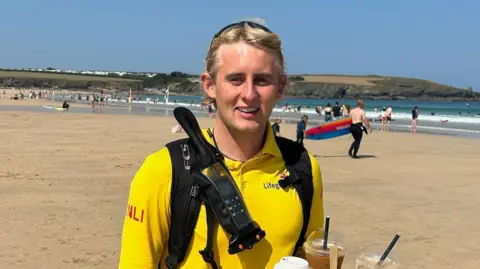 Finlay Maguire A young man with blonde hair is wearing a yellow RNLI lifeguard uniform on a beach on a summer's day. He is smiling into the camera while holding a tray of drinks. 