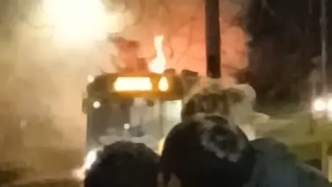 A burry photograph of a bus with flames coming out of its roof, and a number of people standing and watching from the distance. 