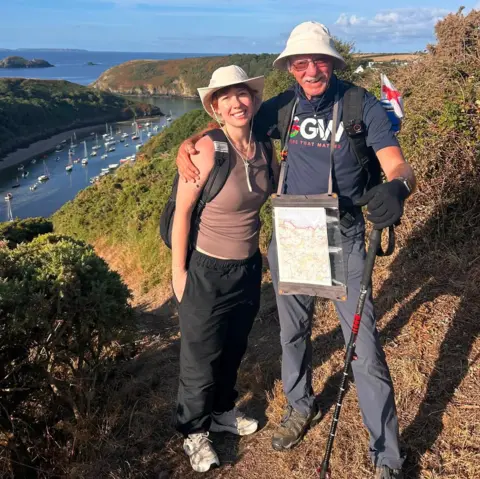 Andy Hockey John Parry and Kyla Norrie stand smiling at the camera. They are both wearing white sun hats, and have their arm around each other's shoulders. John is wearing a 'Grandad's Great Walk' charity t-shirt, while Kyla wears a brown vest. The pair are standing on a footpath in north Pembrokeshire, with a view of hills behind them.