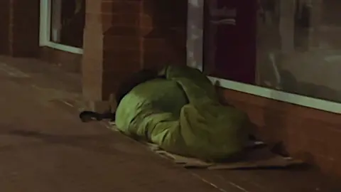 A rough sleeper in a green sleeping bag sleeps beside a row of shop windows 