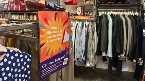 Honor Morgan/BBC The inside of a charity shop which has several rails full of different clothes, including suit jackets, shirts and shoes. There is a sign on the left attached to the end of a rail.