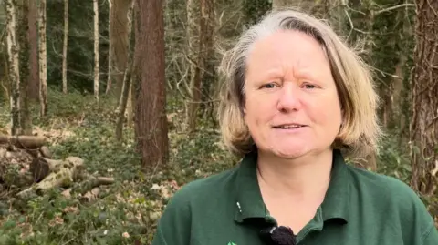 BBC Angela Norwood is pictured with woods behind her. She has light brown, greying hair in a bob and wears a green polo shirt.