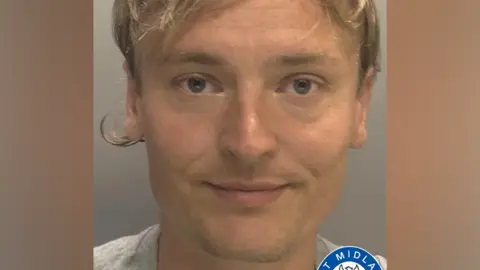A man with brown hair is looking at the camera. Part of the West Midlands Police logo is visible at the bottom of the image.