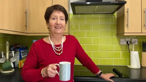 Jan Fuller is smiling at the camera while holding a mug of tea. She is wearing a red knitted top, white necklace and gold watch or bracelet. She has one hand on her kitchen worktop, near the controls of her cooker. To her left are some mugs and breakfast cereals. The kitchen cupboards to her left and right are a light wood effect finish and the tiles on the wall behind her are lime green.