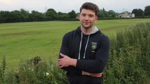 A man - Councillor George Simmonds - standing with his arms folded in front of him wearing a black hoodie and looking straight-faced with a green field in the background. 