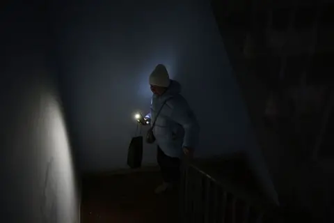 Getty Images A woman, dressed against the cold in woolly hat and think puffer jacket, carries a torch up a dark staircase.