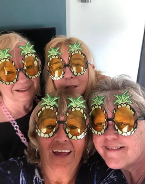 Susie Matthews Four smiling older women pose together for a selfie, each wearing oversized novelty sunglasses shaped like pineapples.