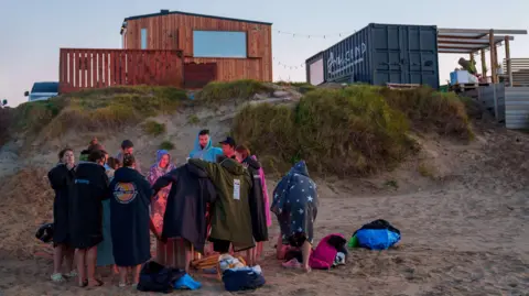 Karl Jones The image shows a group of people on a beach surrounded by a fire, there's a wood fire sauna in the background.