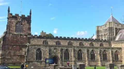 An exterior view of Bangor Cathedral, it is a clear day and there are two people in the foreground of the picture, one walking towards the front door of the cathedral.