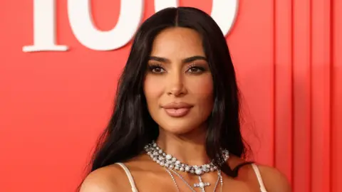 Reuters A headshot of Kim Kardashian as she looks to the left of the camera. She wears multiple necklaces and stands against an orange background.